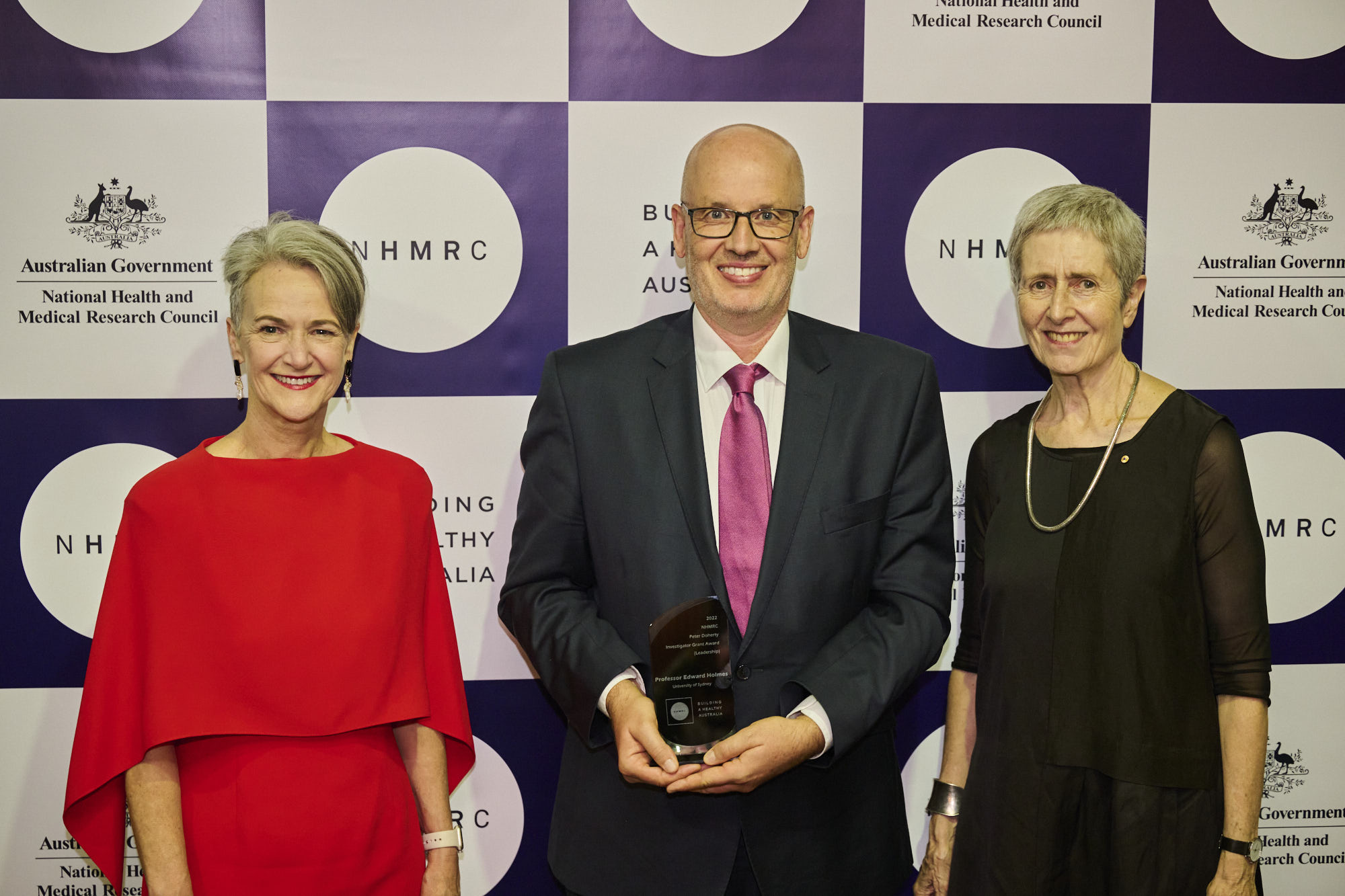 Three people standing in front of media wall with the centre man holding an award.