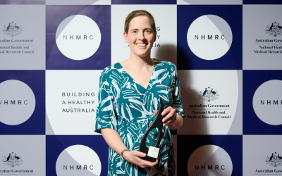 Dr Johanna Birrell holding Research Excellence Award in front of NHMRC branded background