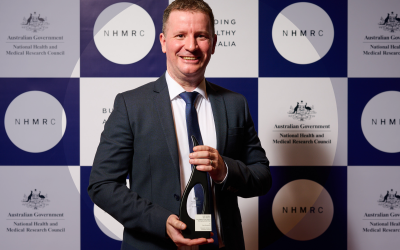 Dr Sean Humphrey holding Research Excellence Award in front of NHMRC branded background