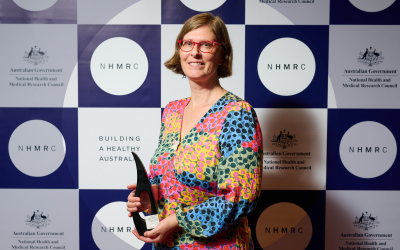 Professor Katy Bell holding research excellence award in front of NHMRC background